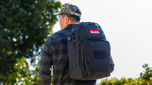 Person wearing a black backpack with a visible brand logo, standing outdoors with trees in the background.